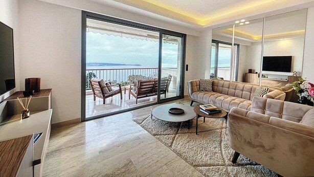 A modern living room with a sofa, coffee table, and a TV. There is a balcony with a view of the sea.