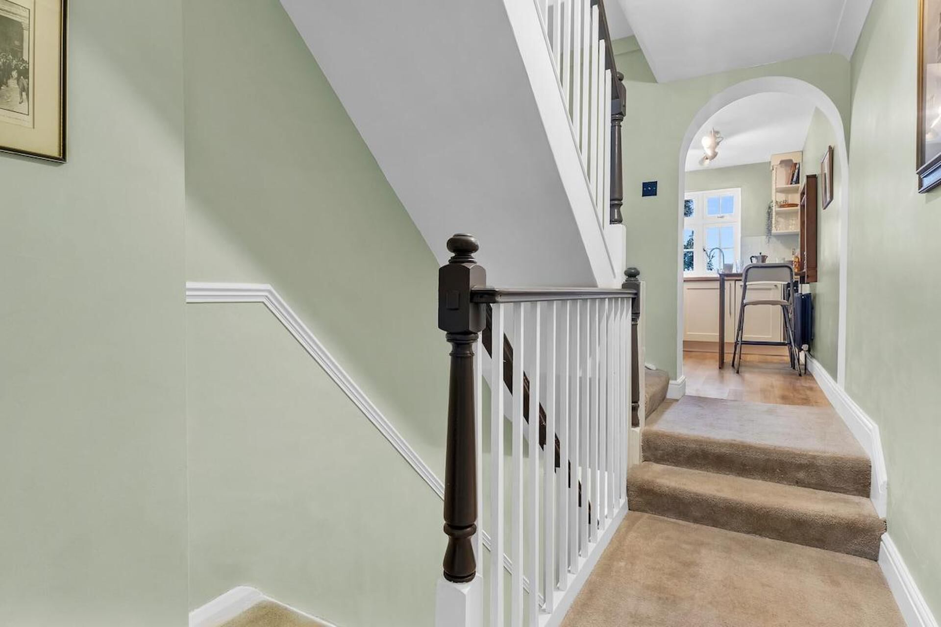 Una escalera con barandillas blancas y un balaustre oscuro, que lleva a una zona de cocina con una ventana y una silla.
