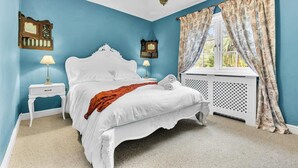 Quarto com cama branca, duas mesas de cabeceira, radiador e janela com cortinas.