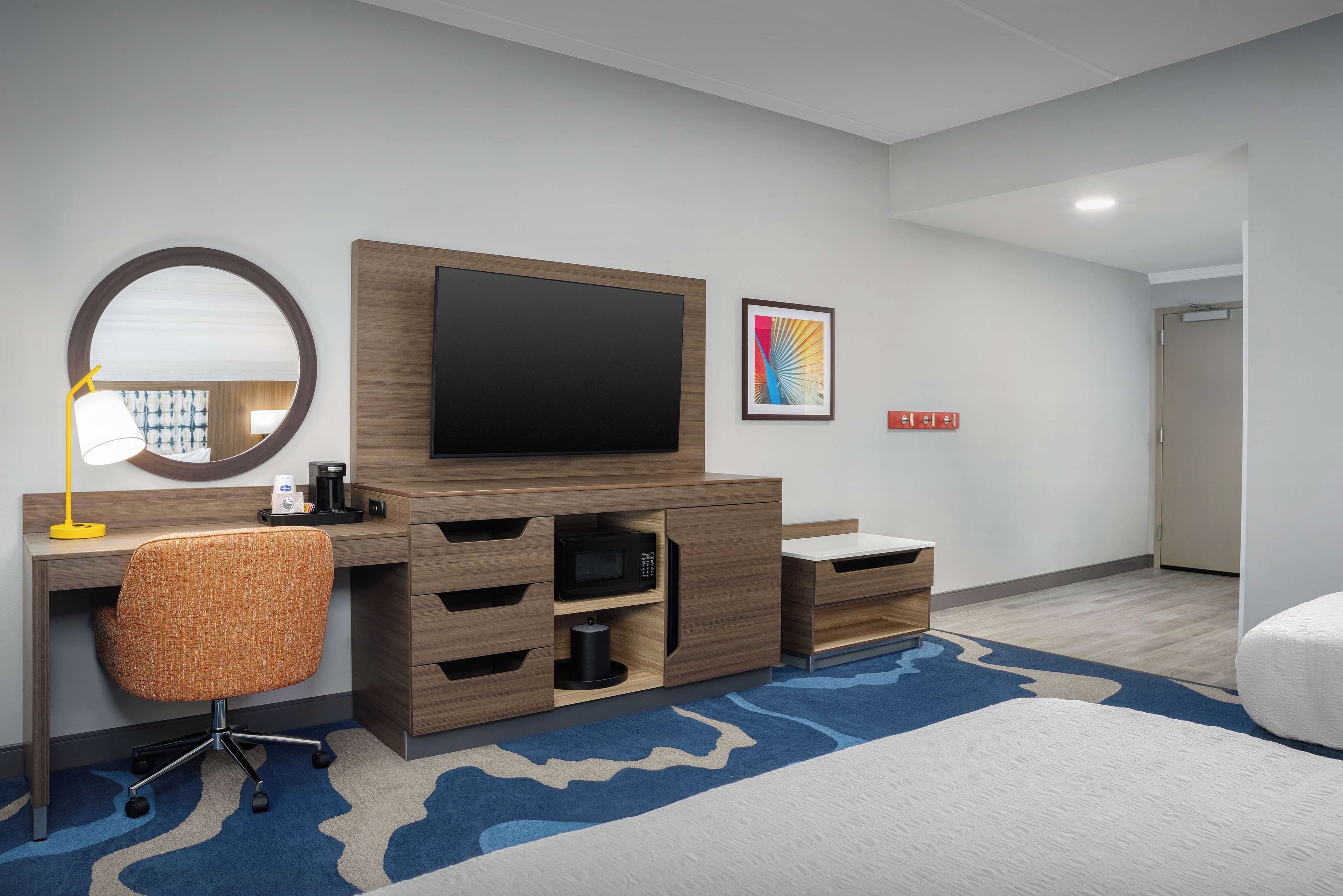Habitación de hotel con un escritorio de madera, un televisor de pantalla plana, un espejo redondo, un microondas y una cama con un tapete azul y blanco a cuadros.