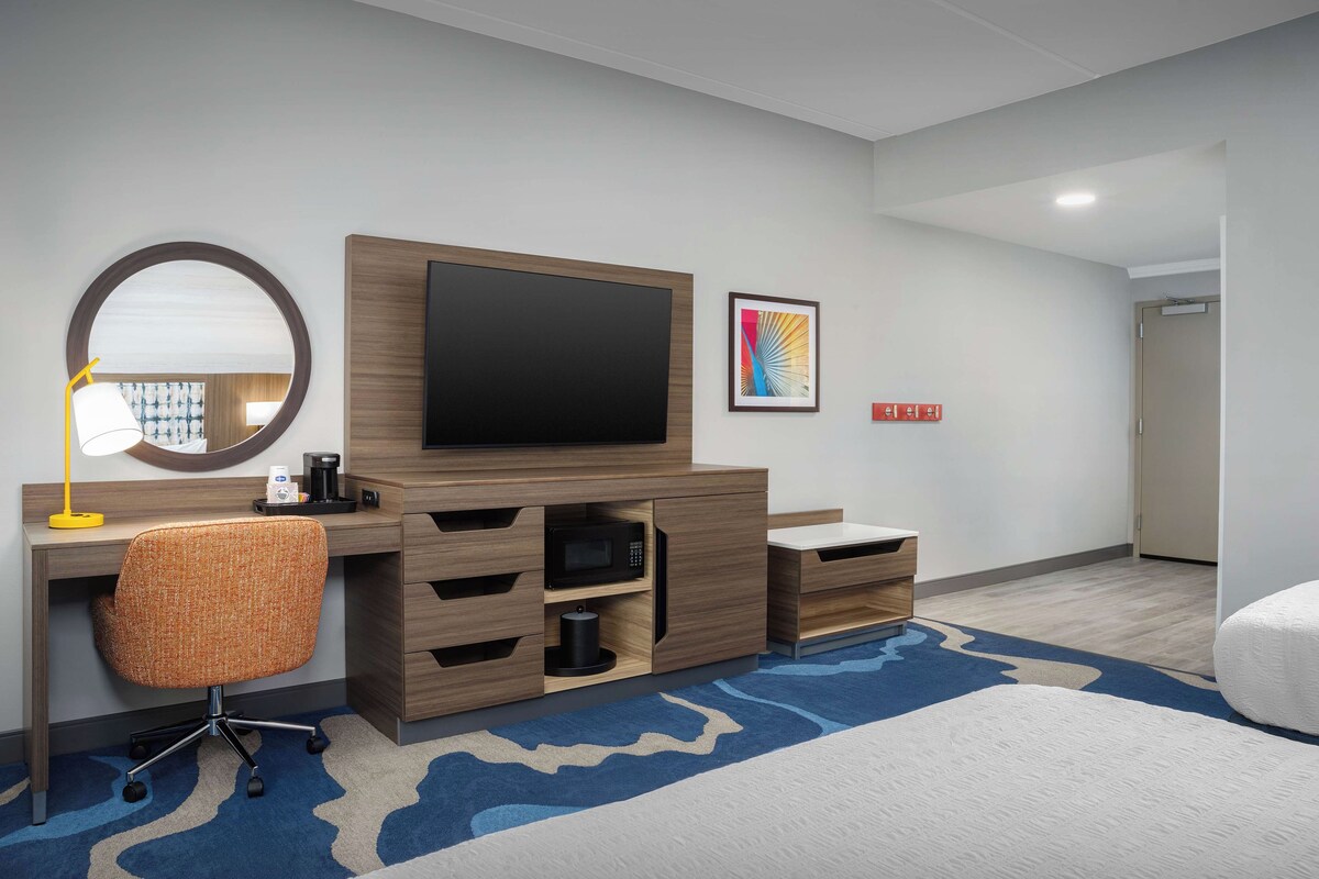 A hotel room with a wooden desk, a flat-screen TV, a round mirror, a microwave, and a bed with a blue and white patterned rug.