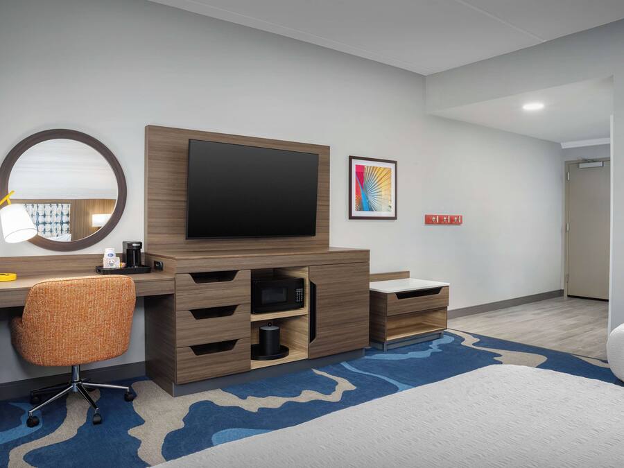 A hotel room with a wooden desk, a flat-screen TV, a round mirror, a microwave, and a bed with a blue and white patterned rug.