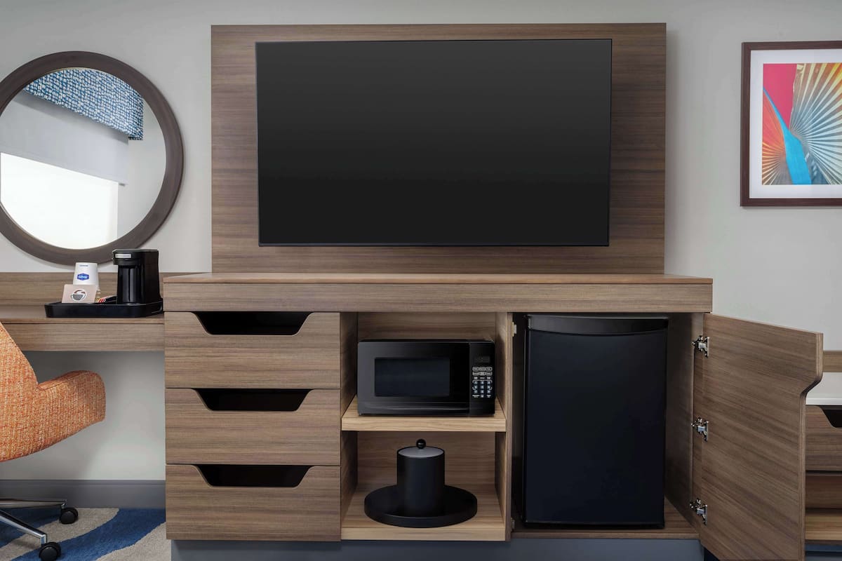 Habitación de hotel moderna con televisión de pantalla plana montada en la pared, microondas, refrigerador y cafetera sobre una consola de madera.
