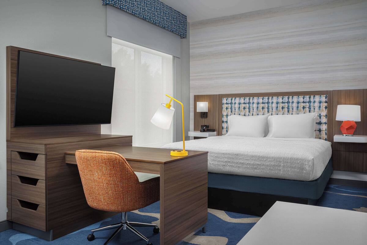 Habitación de hotel con un escritorio de madera, televisión, silla, cama con ropa de cama blanca y mesita de noche con lámpara.