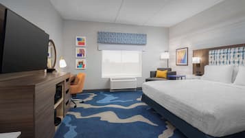 Ein Hotelzimmer mit Bett, Schreibtisch mit Fernseher, Stuhl, Fenster mit Jalousien und Kunstwerken an den Wänden.
