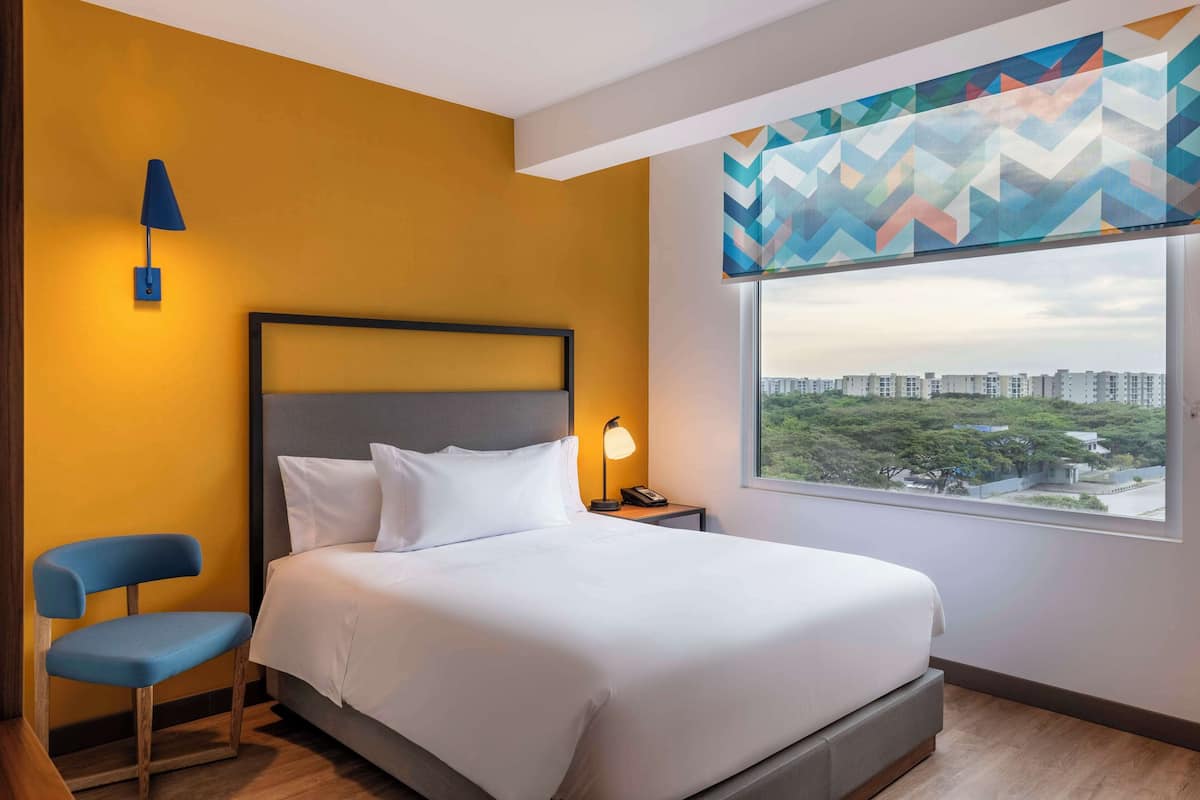 A hotel room with a bed, a chair, a nightstand with a lamp, and a window with a city view.