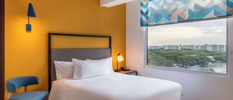 A hotel room with a bed, a chair, a nightstand with a lamp, and a window with a city view.