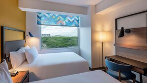 A hotel room with a bed, a desk, a chair, a lamp, and a window with a view of greenery and buildings. - Tru By Hilton Cali Sur (Cali)