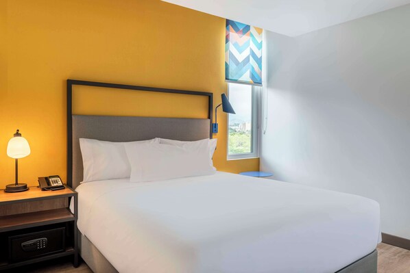 A hotel room with a bed, a nightstand with a lamp and phone, and a window with a view of buildings. - Tru By Hilton Cali Sur (Cali)