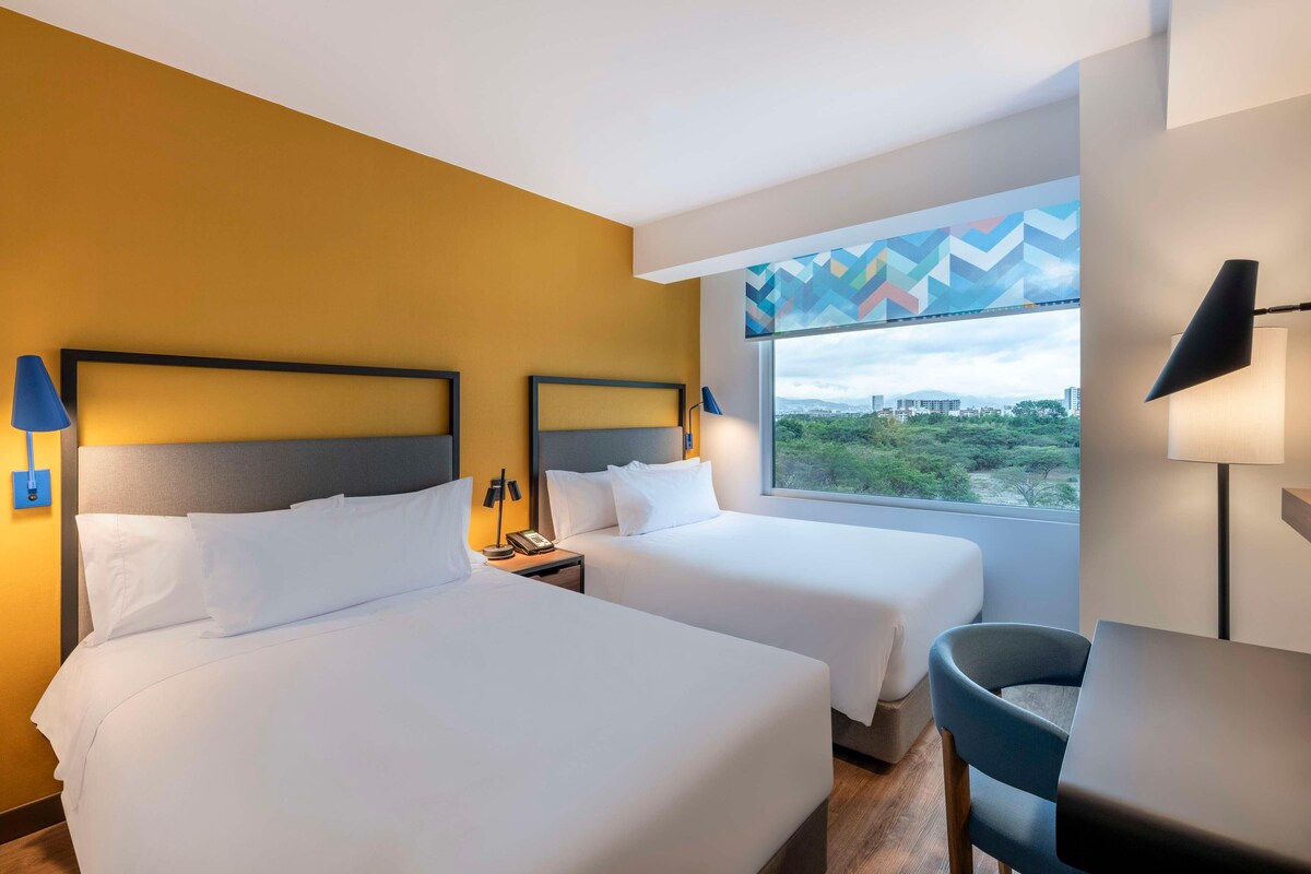 A hotel room with two beds, a desk, and a chair. There is a window with a view of a cityscape and greenery.
