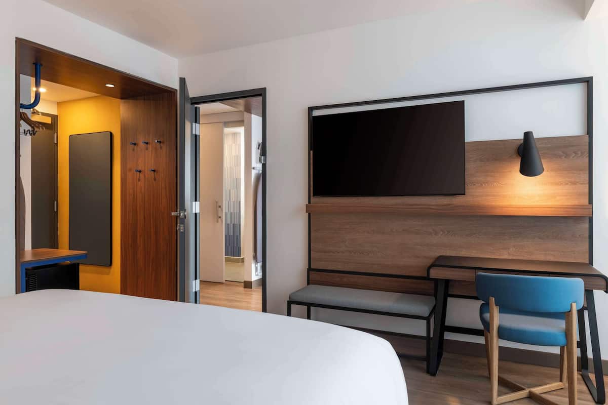 A modern hotel room with a large bed, a desk, a chair, a TV mounted on the wall, and a wooden wardrobe.