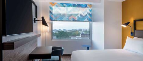 A hotel room with a bed, a desk, a chair, a TV, a window with a view of buildings, and a patterned curtain.
