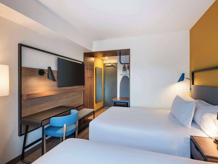 Quarto de hotel com duas camas, uma escrivaninha com cadeira, televisão e espelho.
