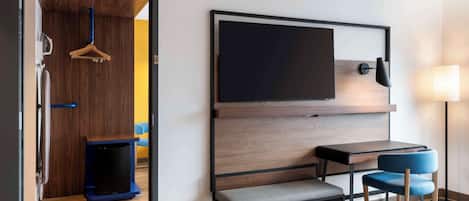 A hotel room with a wooden wardrobe, a flat-screen TV mounted on the wall, a small table with two chairs, and a bedside lamp.