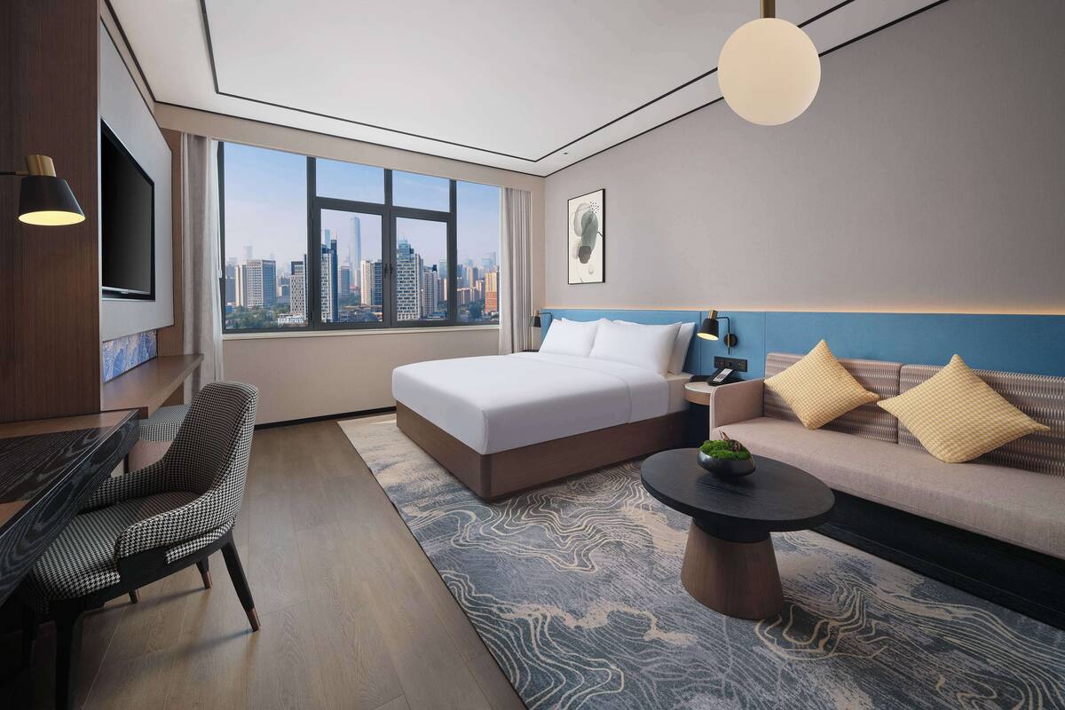 Quarto de hotel moderno com uma cama grande, um sofá, área de refeições e vista para a cidade.