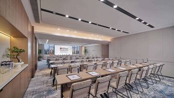 A conference room with long tables and chairs, a large screen displaying Hilton Garden Inn, and a city view through large windows.