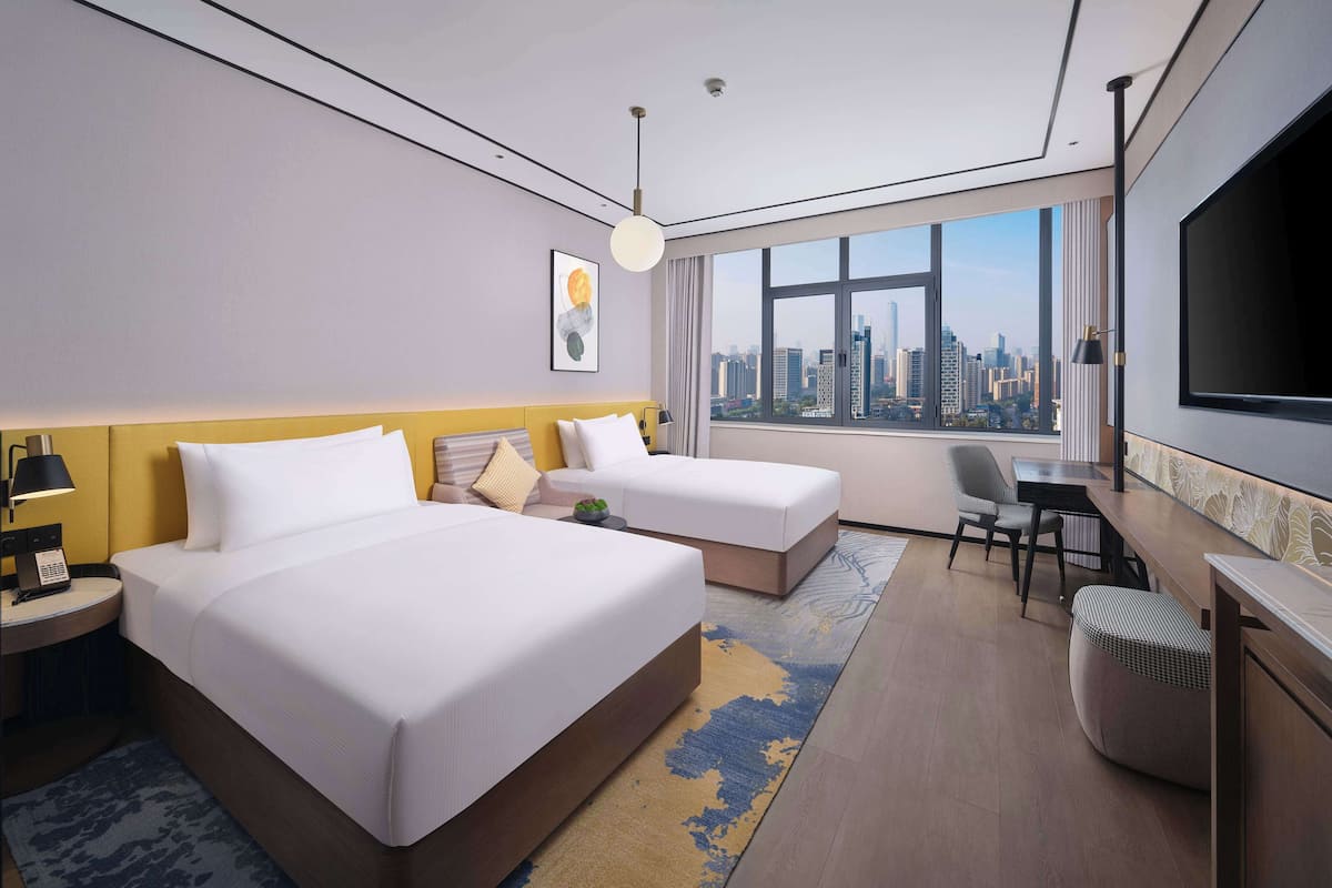 Quarto de hotel com duas camas, uma mesa, uma cadeira, televisão e uma janela grande com vista para a cidade.