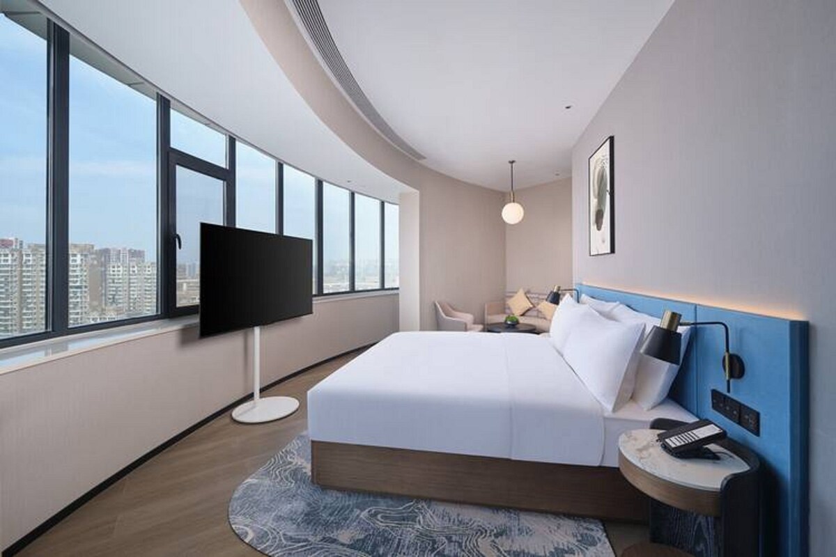 A modern hotel room with a large bed, a flat-screen TV, a bedside table with a lamp, and a view of the city through large windows.