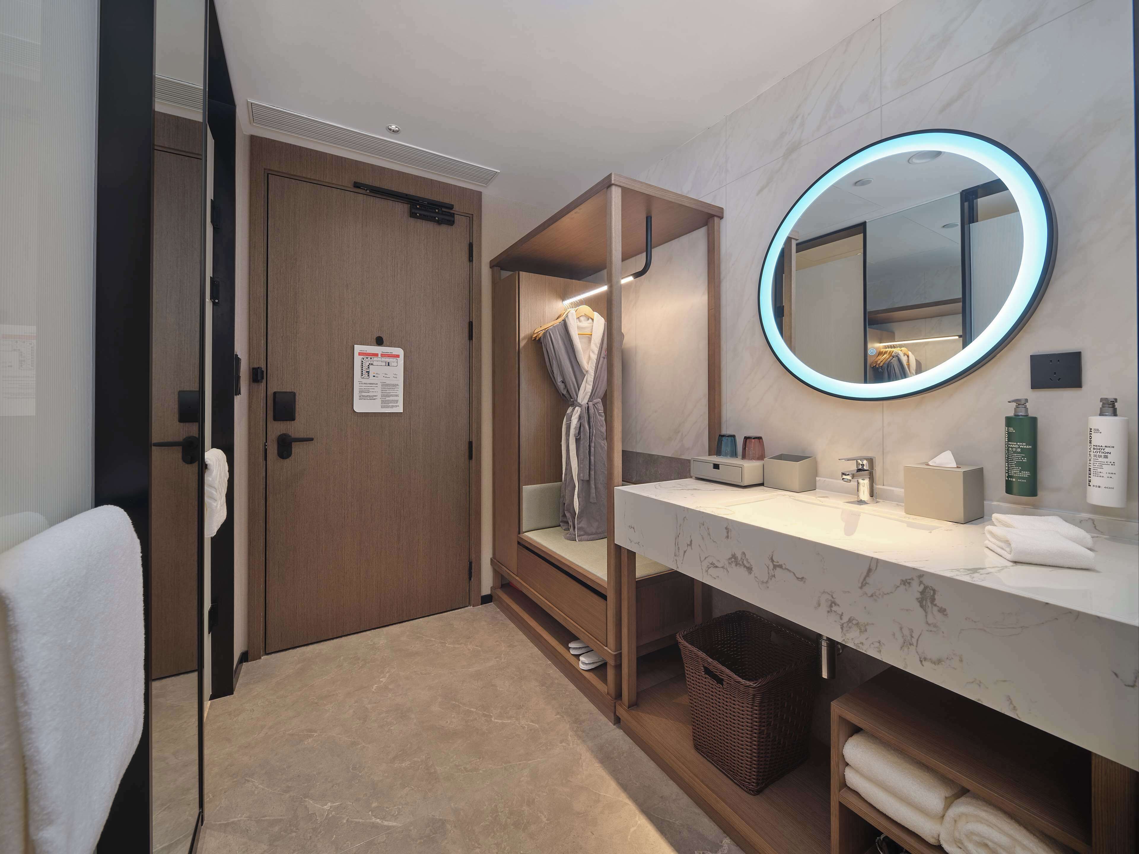 Un baño moderno con puerta de madera, espejo con luz circular, tocador con encimera de mármol y toallero.