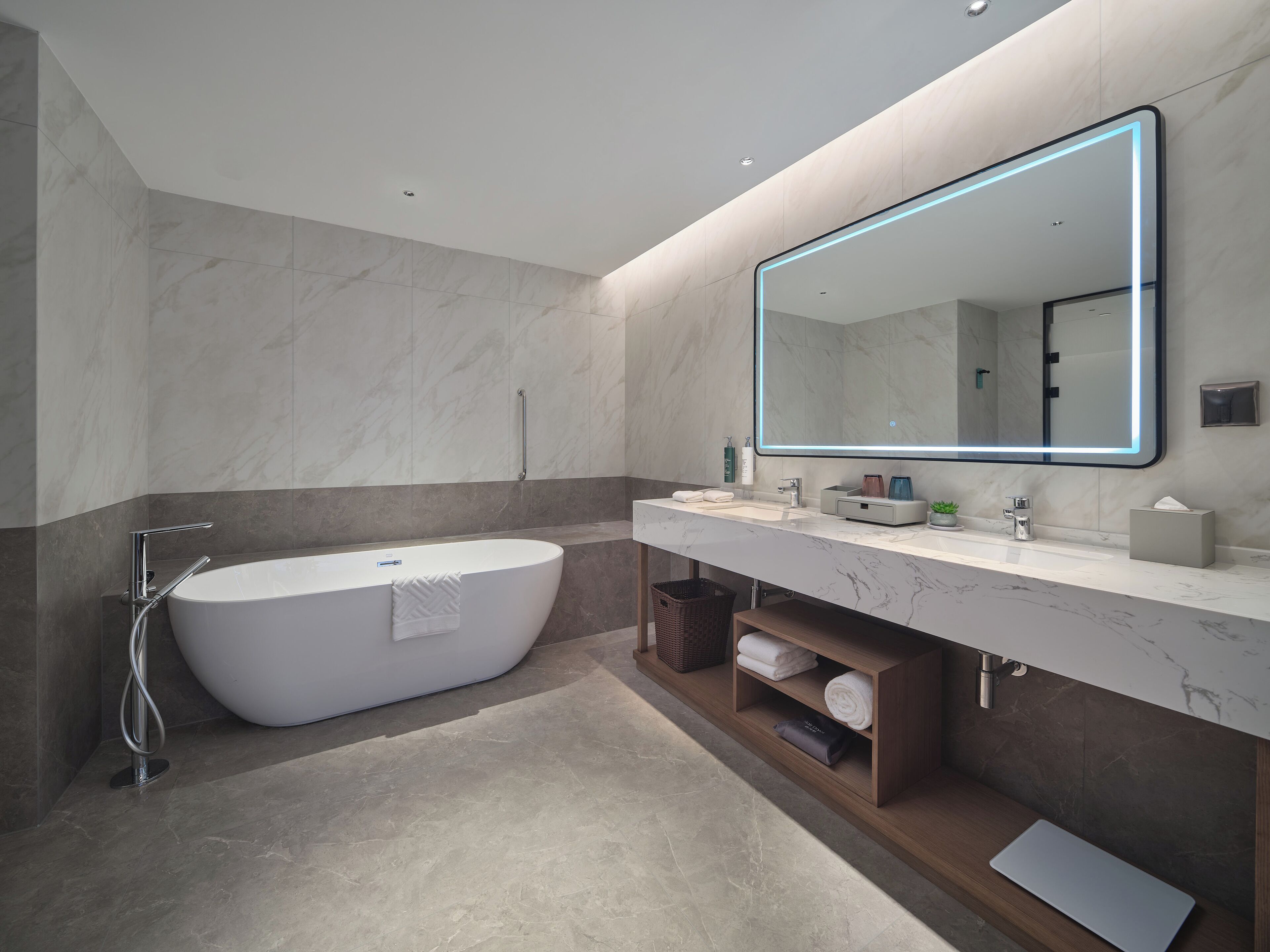 A modern bathroom with a freestanding bathtub, a large mirror with LED lighting, a vanity with a marble countertop, and a towel rack.