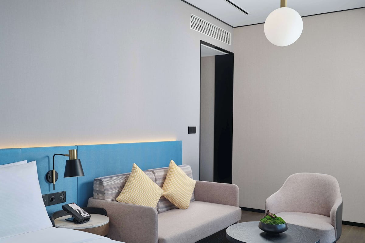 Quarto de hotel moderno com uma parede azul de destaque, um sofá com almofadas estampadas, uma mesa redonda com uma planta e um abajur.