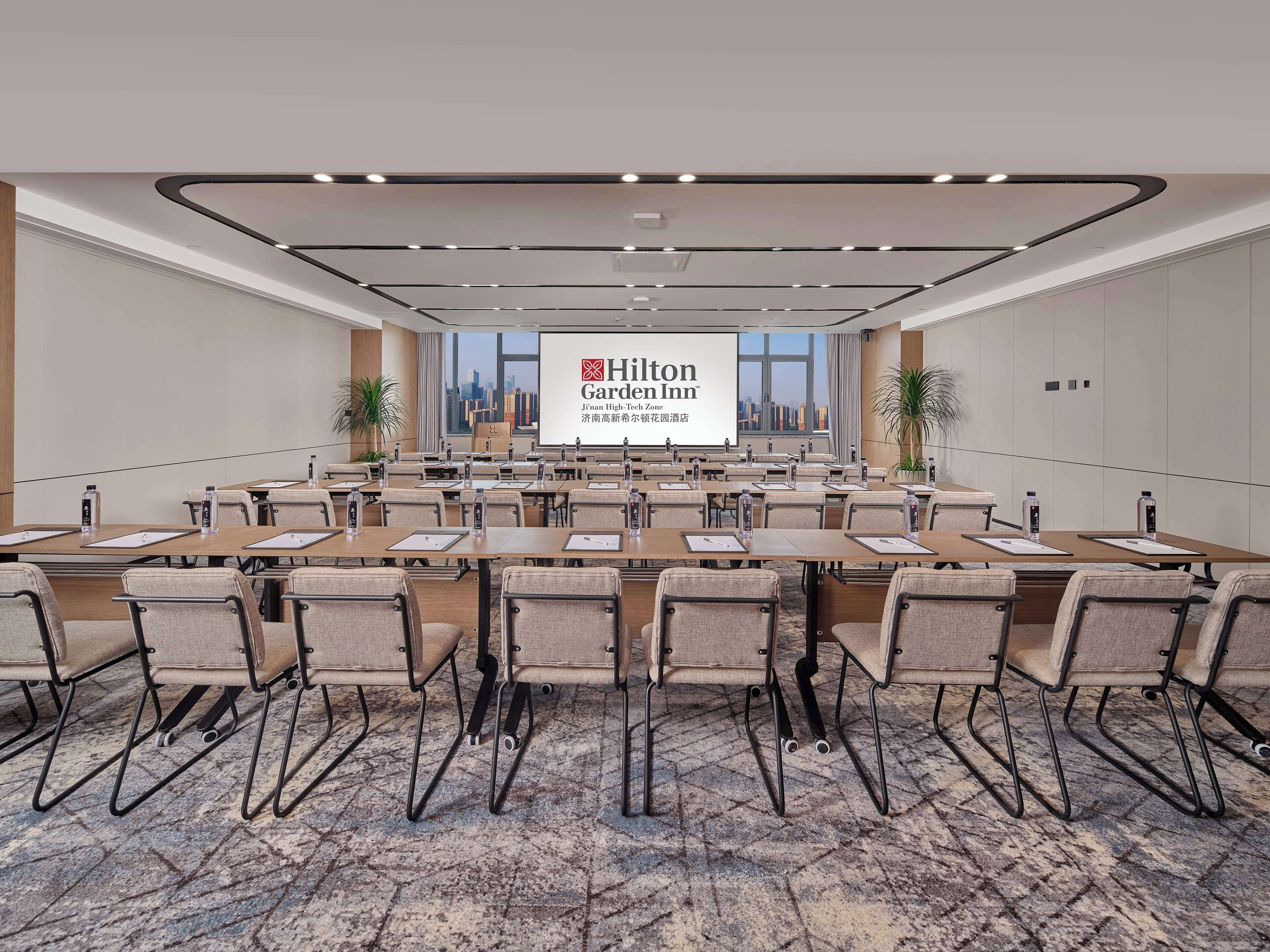 A conference room with a large screen displaying the Hilton Garden Inn logo, multiple tables with chairs, and a city view through the windows.