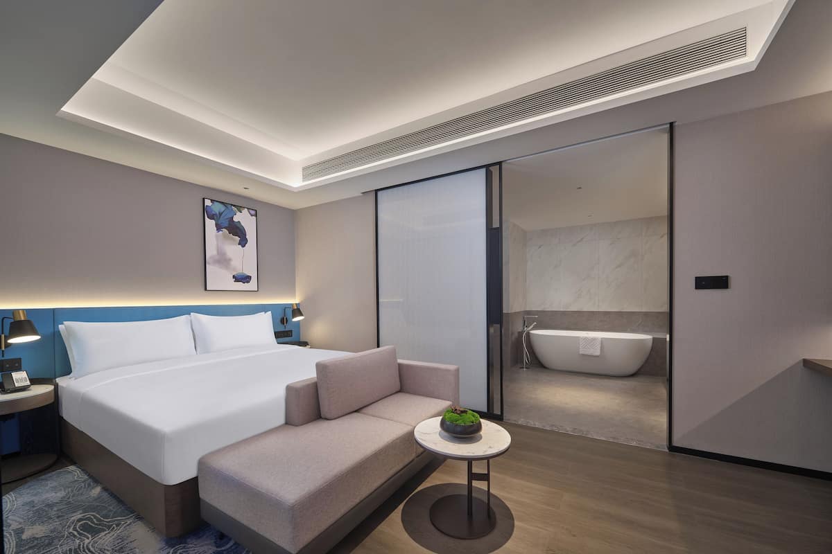 Quarto de hotel moderno com uma cama grande, um sofá pequeno, uma mesa redonda com uma planta e um banheiro separado com banheira.