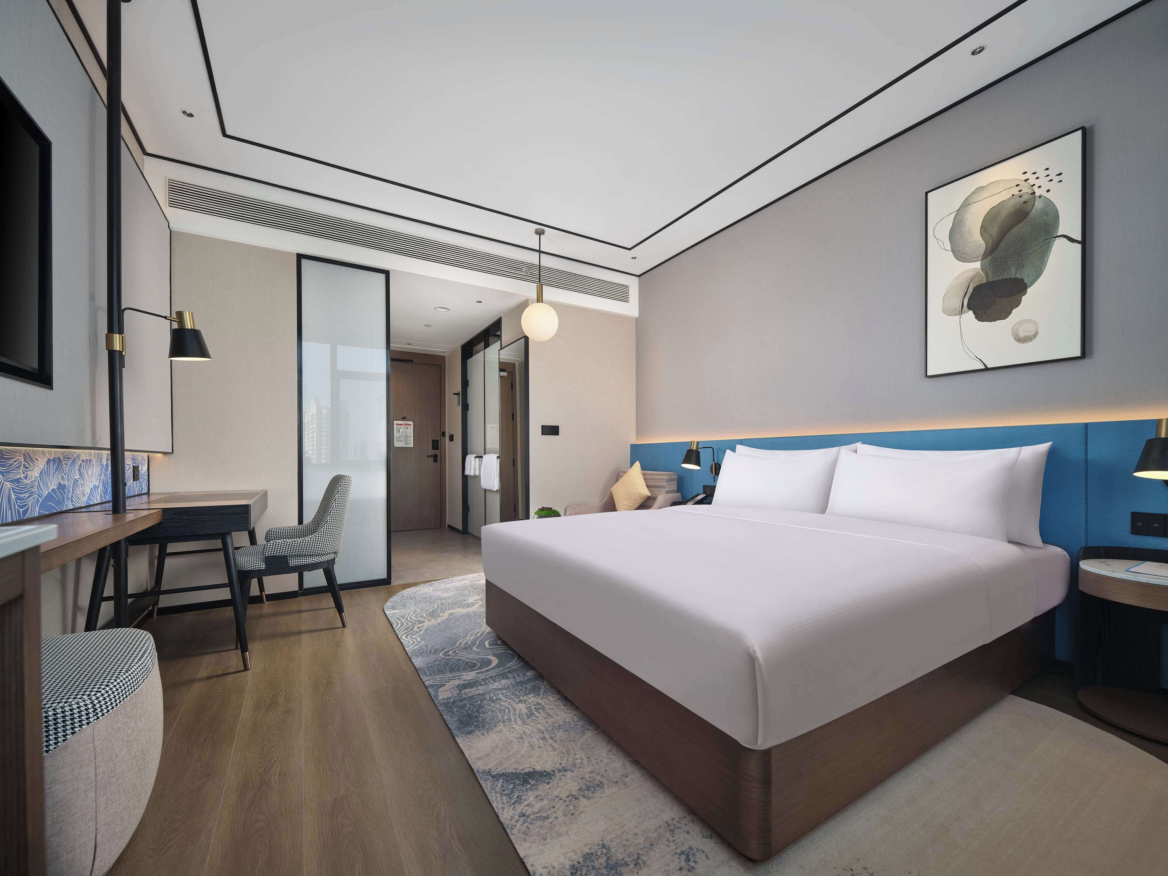 A modern hotel room with a large bed, a desk, and a chair. There is a painting on the wall and a lamp on the nightstand.