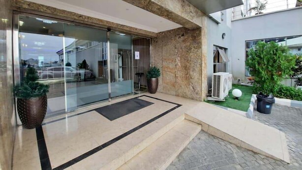 A modern building entrance with a glass door, a small outdoor seating area with a potted plant, and a paved pathway leading to the entrance. - The Patron Hotel Suites Lekki (Lekki)