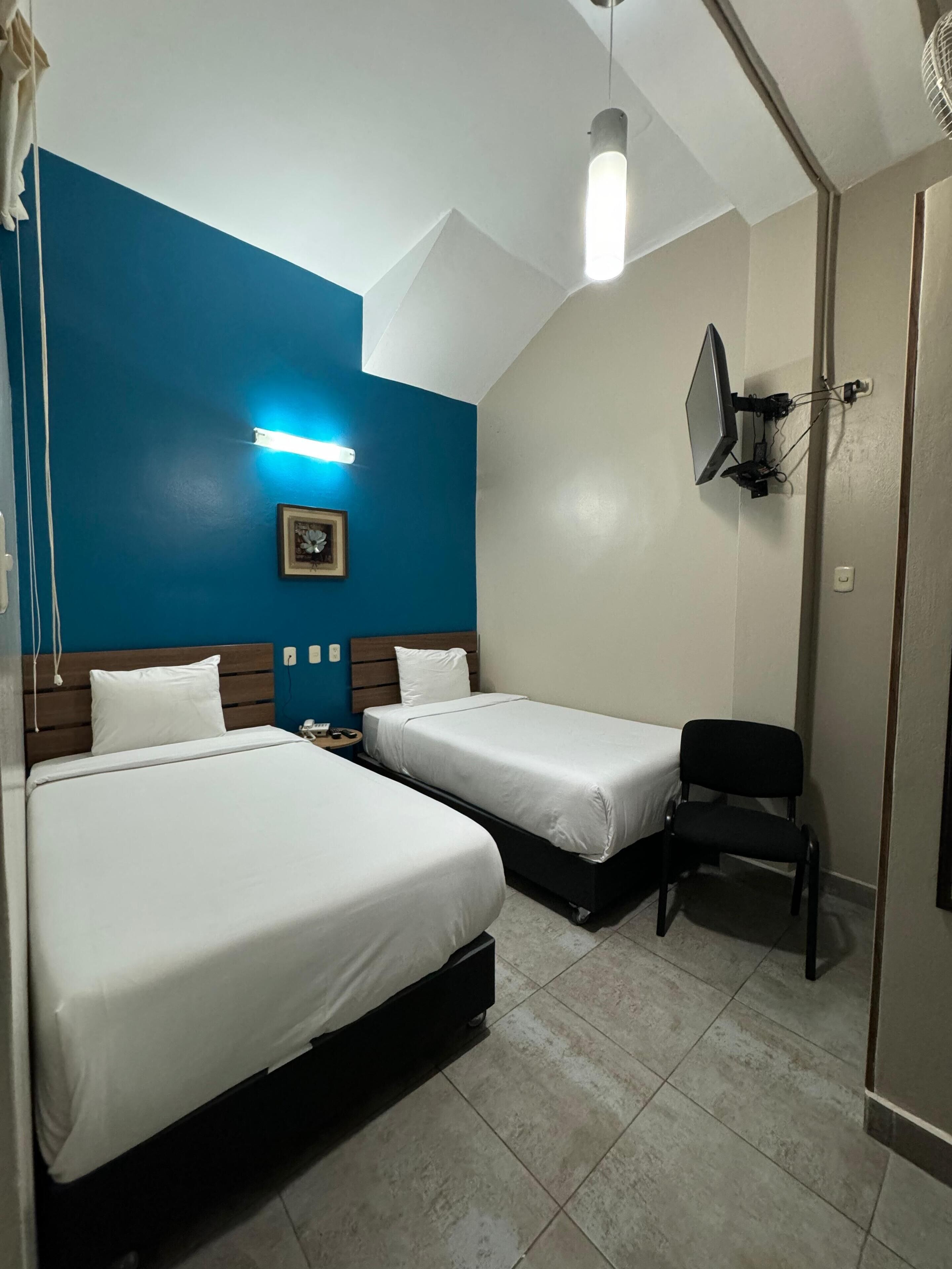 A hotel room with two beds, a desk, a chair, a ceiling fan, and a wall-mounted TV.