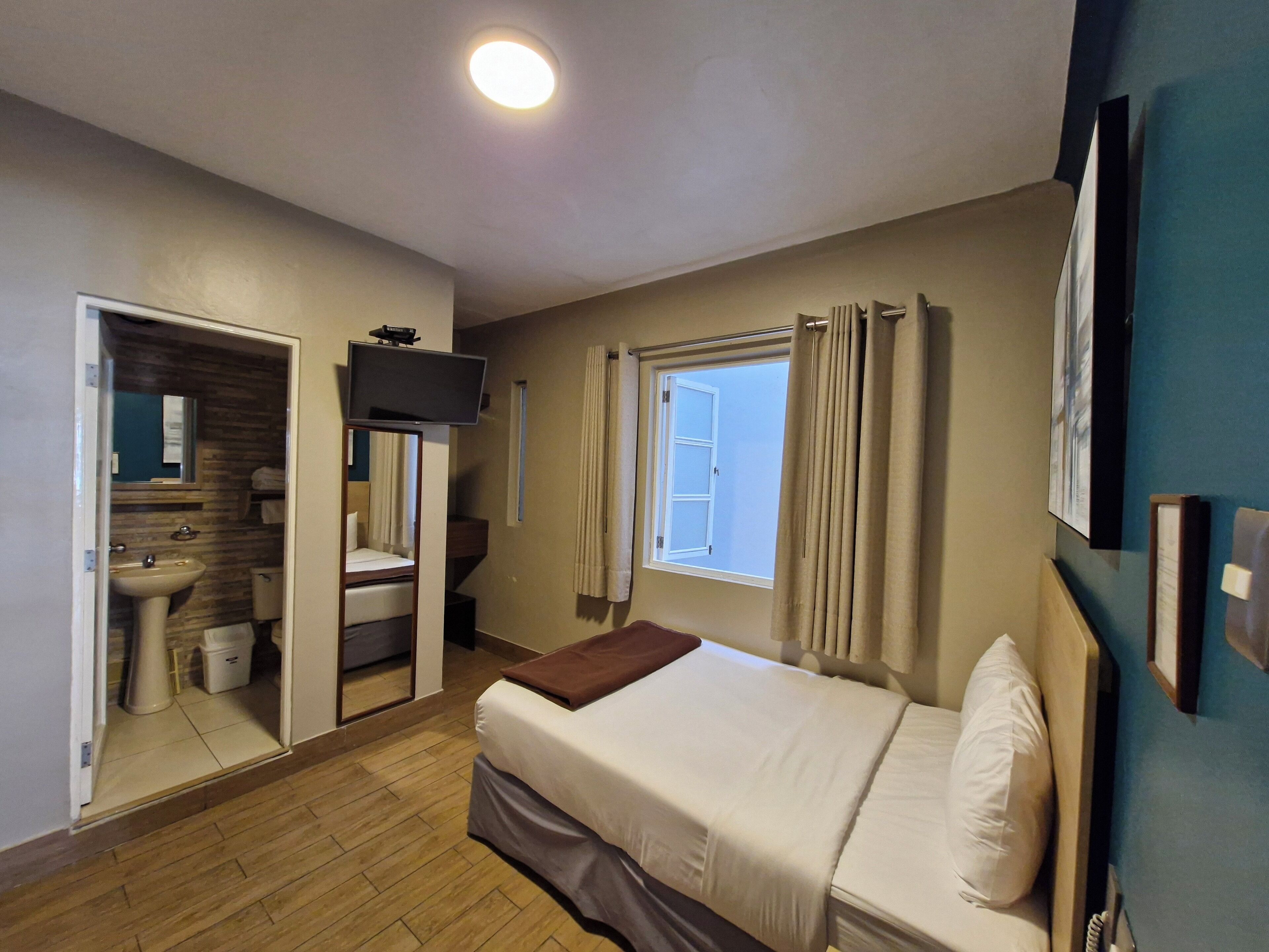 A bedroom with a bed, a TV mounted on the wall, a window with curtains, and a bathroom visible through an open door.