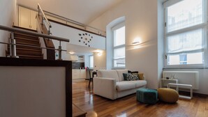 A modern apartment with a staircase, a living area with a sofa and ottoman, a kitchen in the background, and large windows providing natural light. - Il Colibrì by Wonderful Italy (Turin)