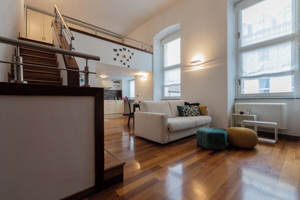 A modern apartment with a staircase, a living area with a sofa and ottoman, a kitchen in the background, and large windows providing natural light.