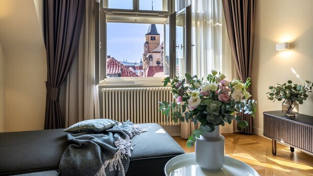 Una habitación con un ventanal amplio, vista a una torre, un sofá con una manta y un florero con flores.