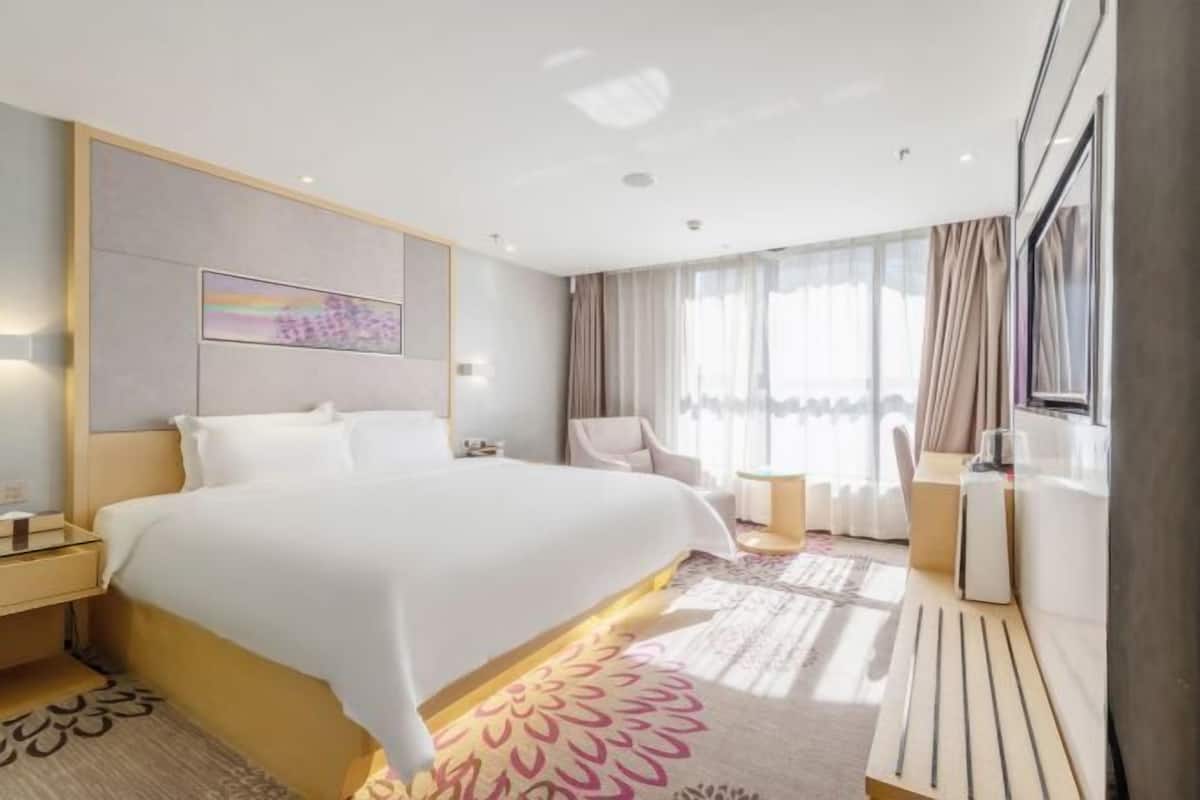 A modern hotel room with a large bed, a bedside table, a chair, a desk, and a large window with curtains.