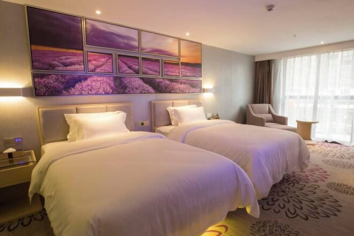 A hotel room with two beds, a large window with curtains, and a wall-mounted artwork.