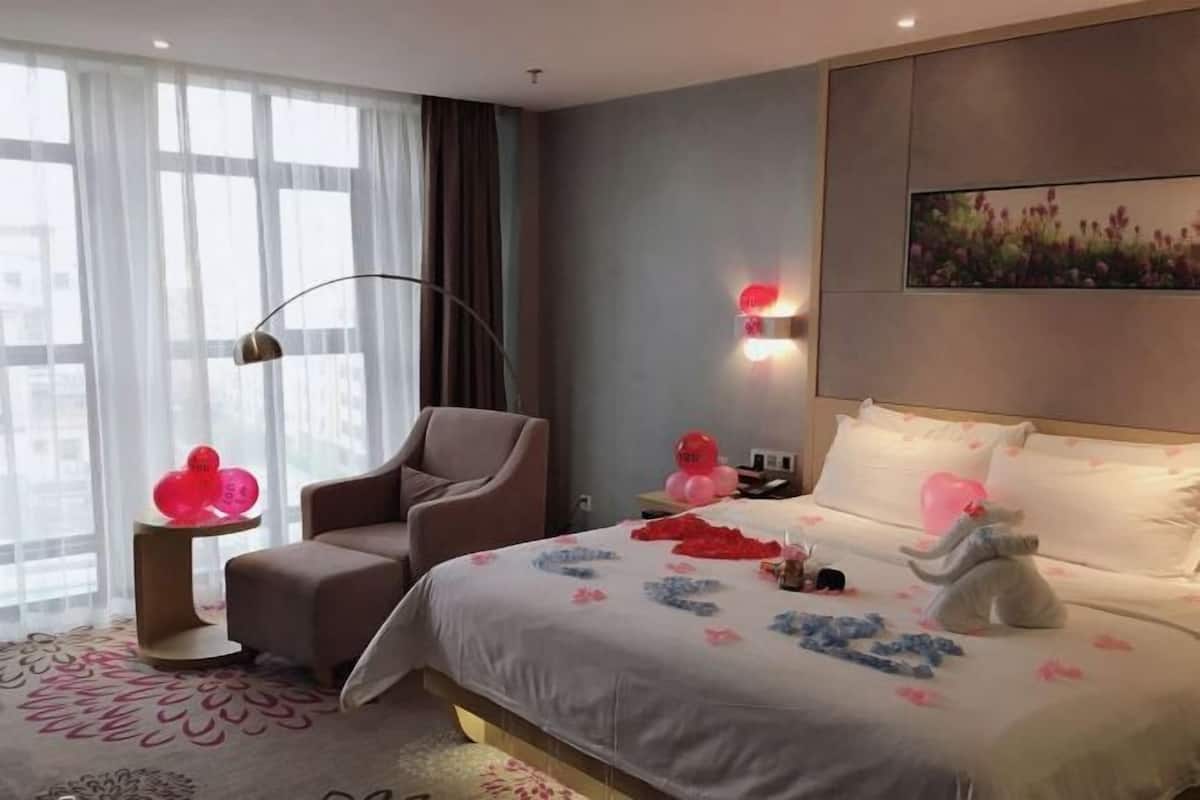 A hotel room with a bed, a chair, a side table with balloons, and a painting on the wall.