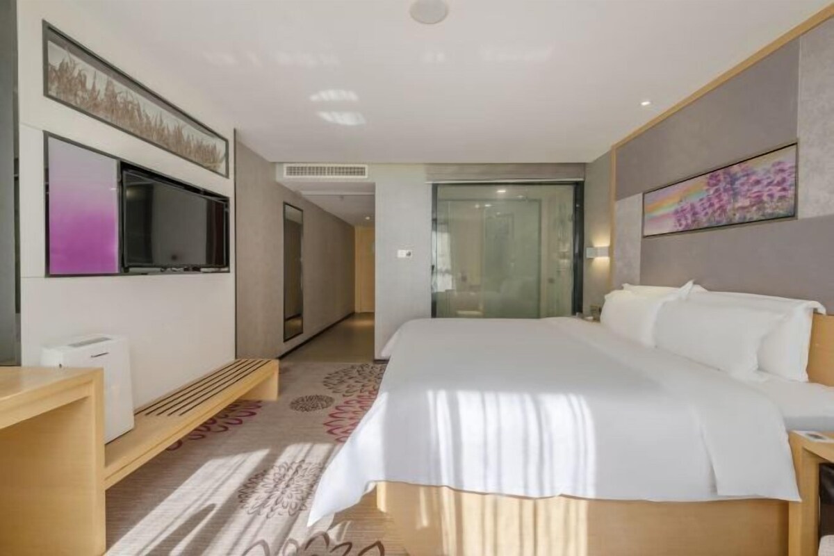 A modern hotel room with a large bed, a flat-screen TV, a bedside table, and a glass door leading to a hallway.