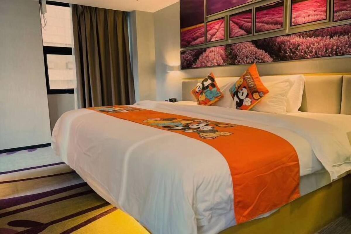 A hotel room with a bed, a window with curtains, a wall with framed pictures, and a bedspread with cartoon characters.