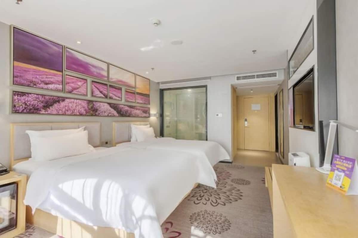A hotel room with two beds, a large window, a wall-mounted artwork, and a desk with a chair.