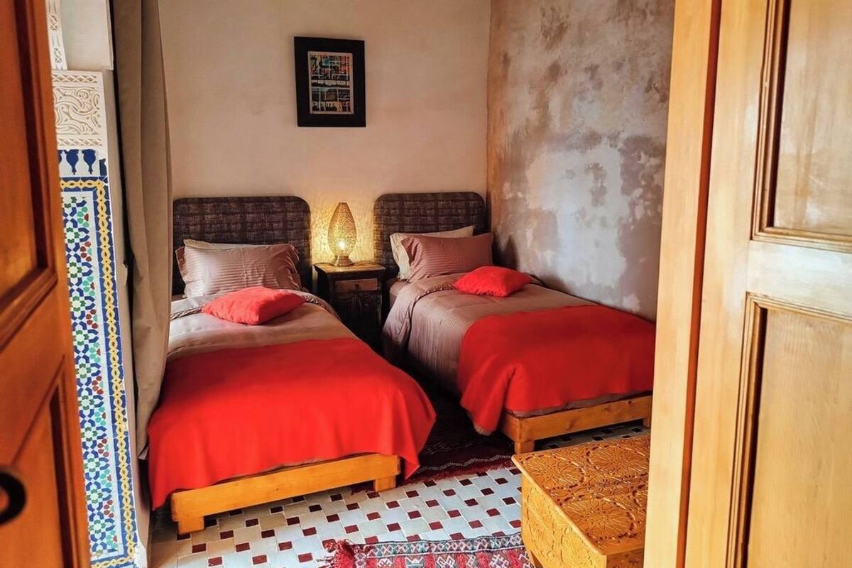 A room with two beds, each with a red blanket and pillows, a nightstand with a lamp, and a patterned rug on the floor.