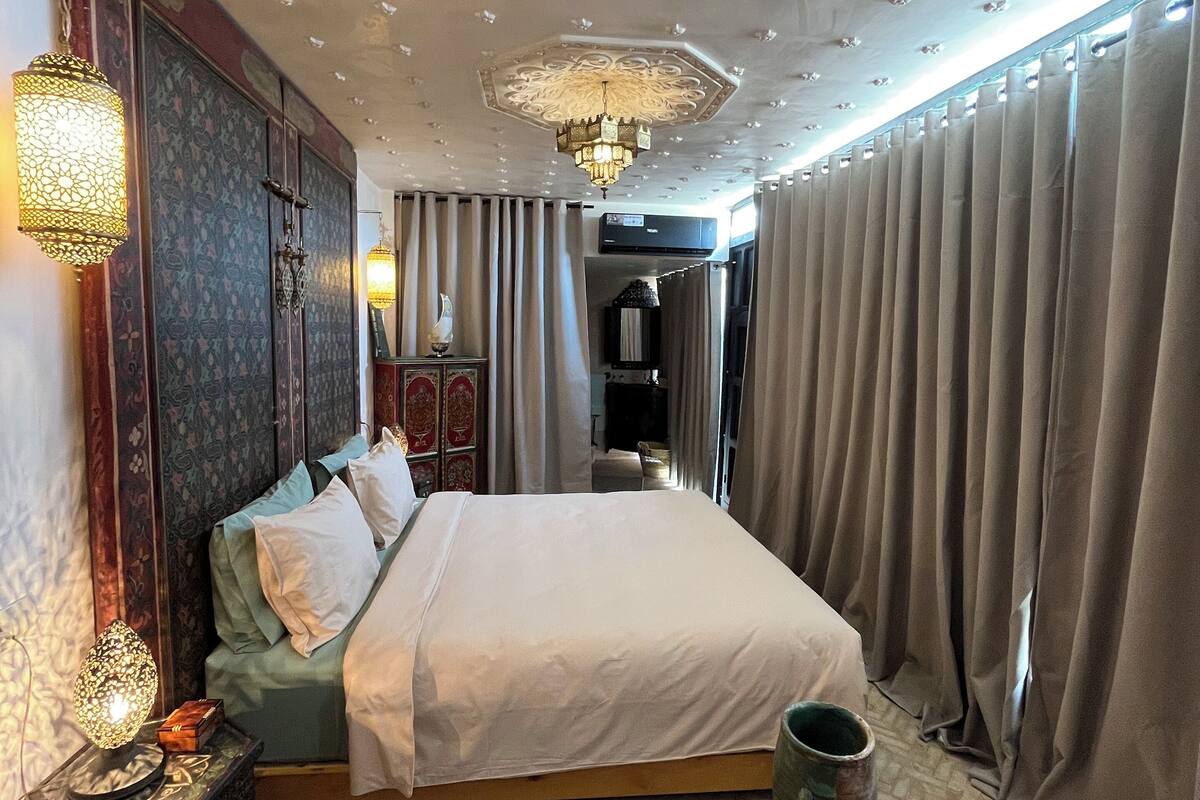 A hotel room with a large bed, a chandelier, a decorative wall, and a view of the hallway.
