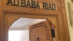 En trädörr med skylten ”ALIBABA RIAD” ovanför, som leder in till en utomhusplats med sittplatser, ett mönstrat golv och en hängstol.