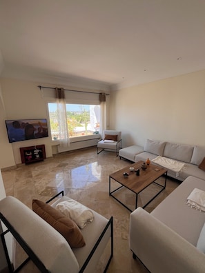 A living room with a sofa, armchairs, a coffee table, a TV, and a window with curtains.