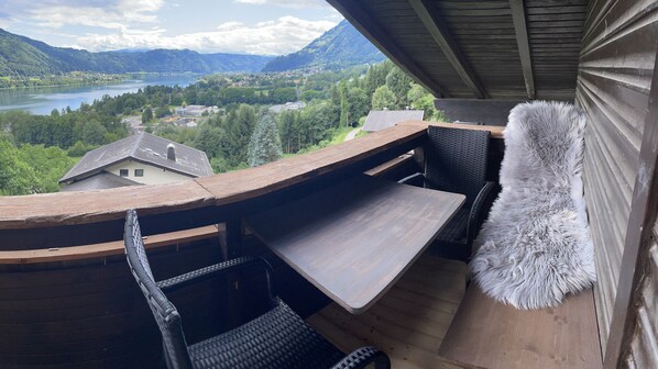 A balcony with a wooden table and chairs, overlooking a scenic view of a lake and mountains.