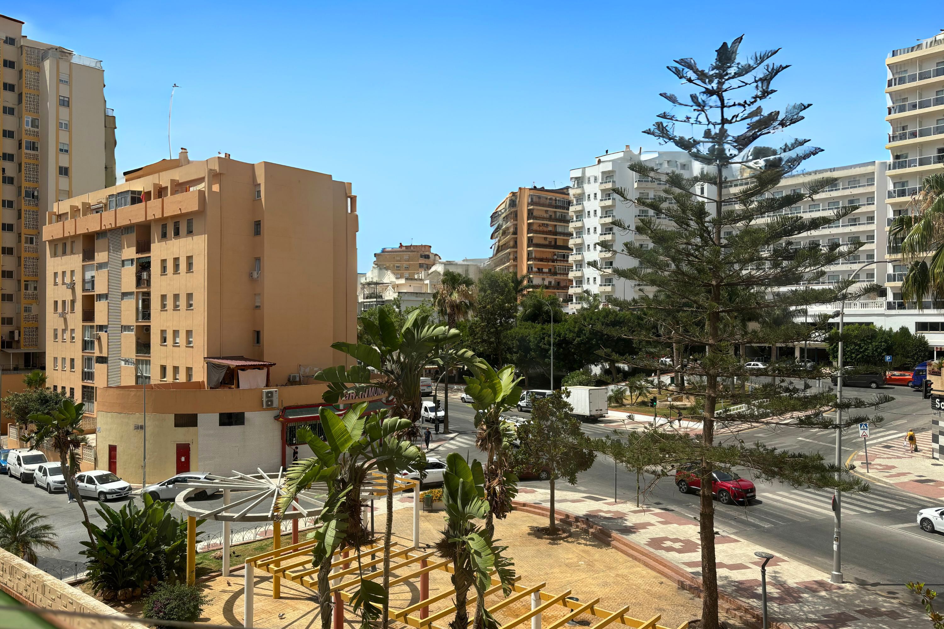 A multi-story building with balconies, a parking area with cars, and a street with palm trees and a pedestrian crossing.