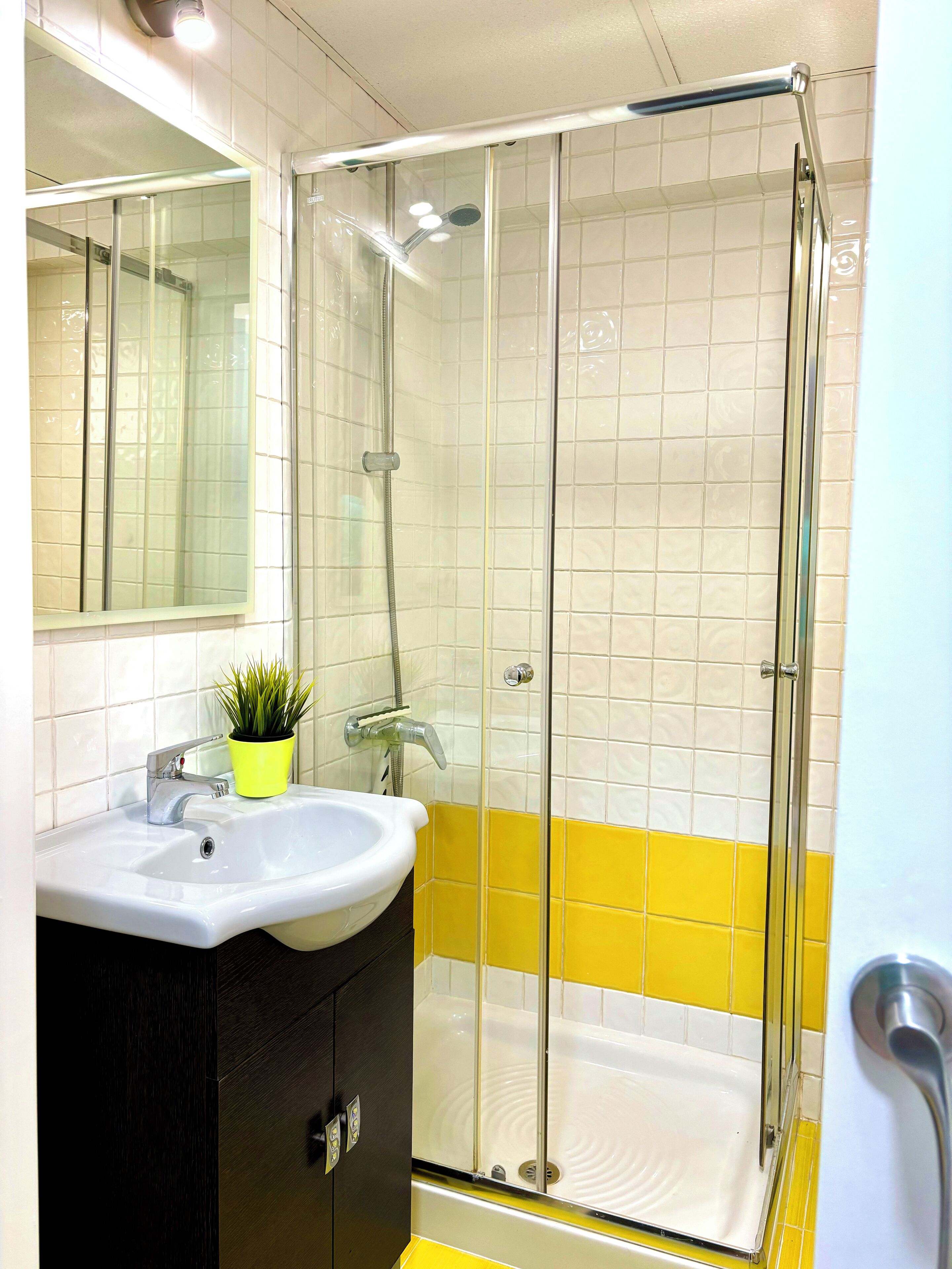 A bathroom with a glass-enclosed shower, a sink with a mirror, and yellow and white tiled walls.