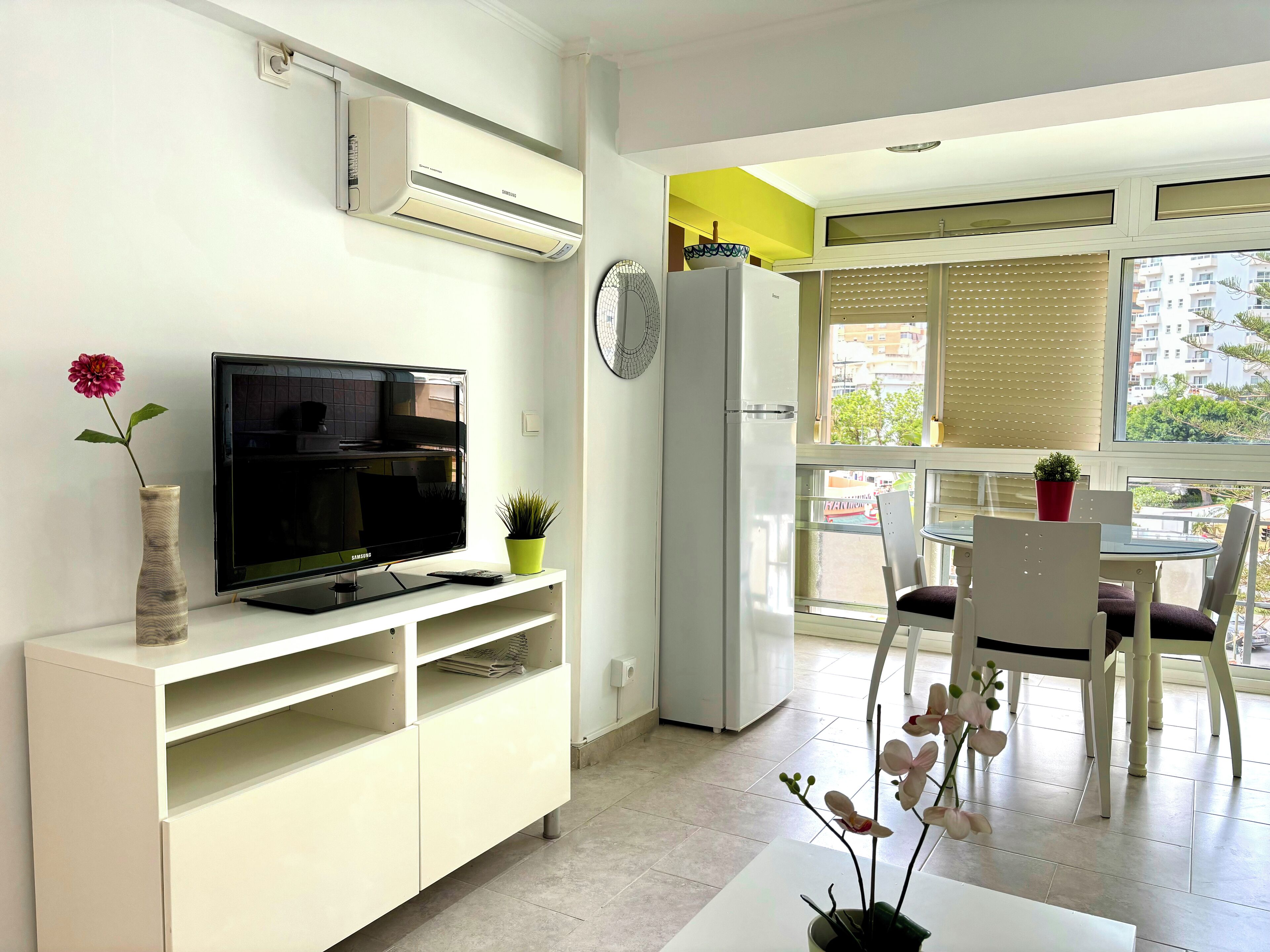 Una sala moderna con un televisor de pantalla plana, muebles blancos, una zona de comedor con mesa y sillas, y un ventanal con persianas.