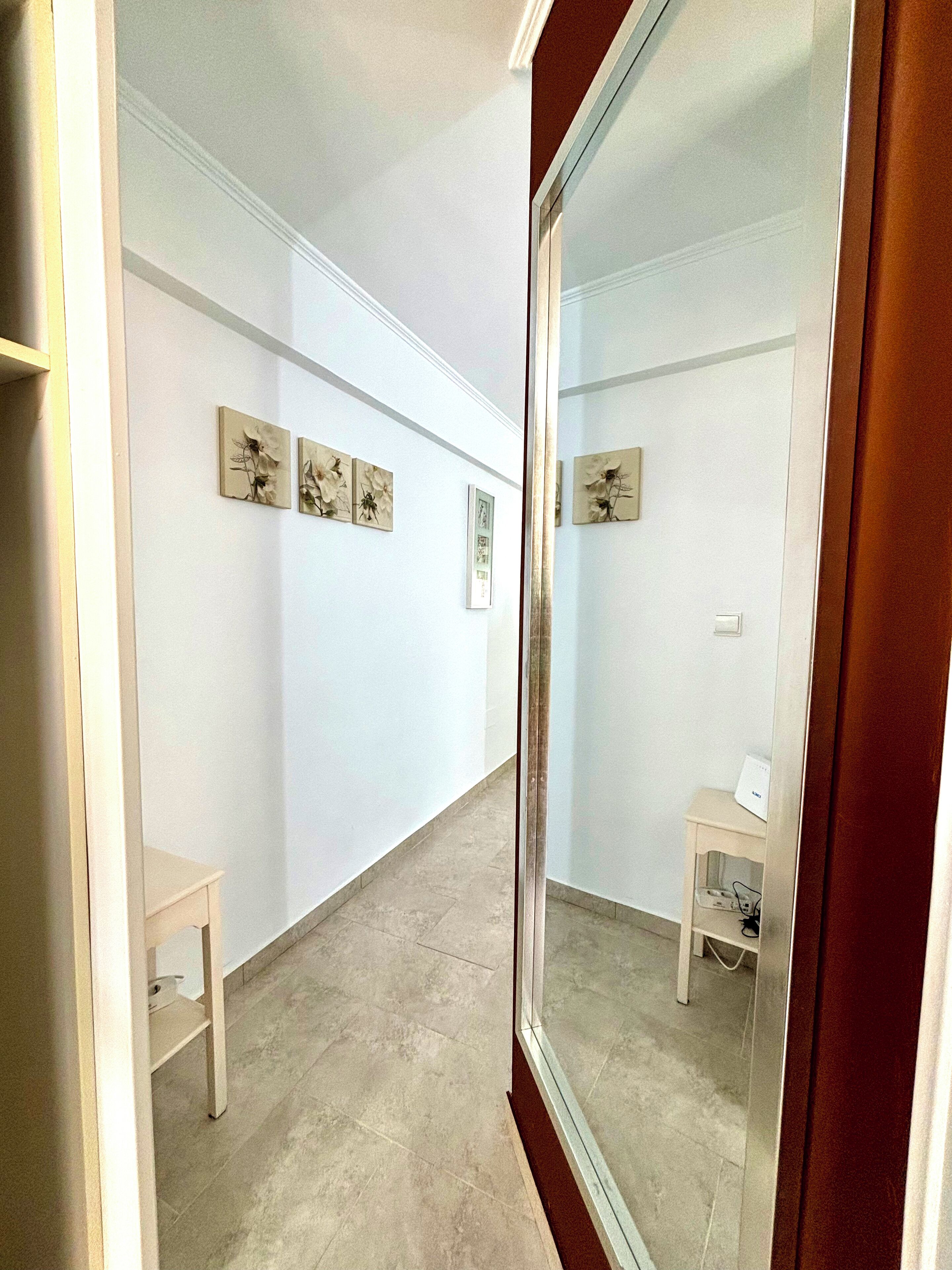 Un pasillo con un espejo que refleja una habitación, una mesita pequeña y obras de arte en las paredes.