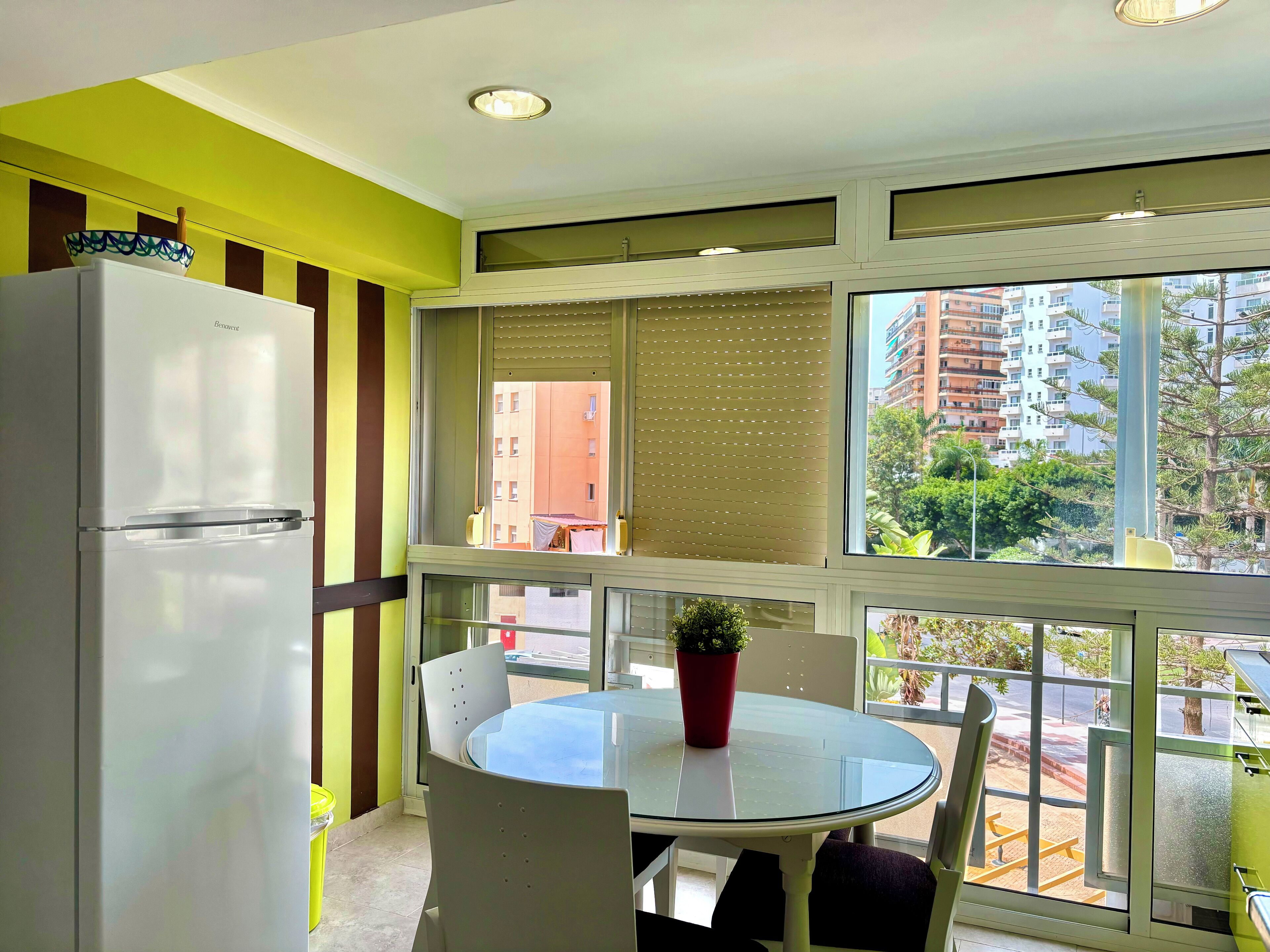 Cocina compacta con refrigerador, mesa de comedor con sillas y una ventana con persianas.